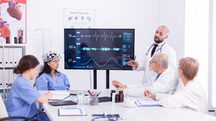 Doctor discussing about the future of neurology with medical staff in hospital meeting room. Monitor shows modern brain study while team of scientist adjusts the device.